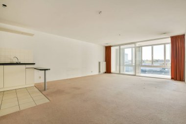 Geniş oturma odasında geniş pencereler, bej halı ve parlak ve havadar bir atmosfer sağlayan minimalist mutfak düzeneği bulunur..