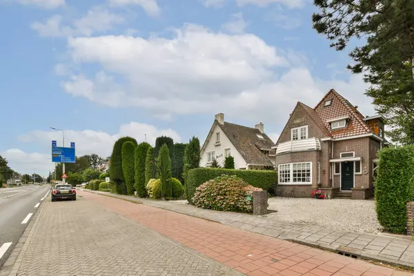 Bereketli yeşillik ve bakımlı çalılarla çevrili büyüleyici evlerin yer aldığı sakin bir yol kenarı manzarası, pitoresk bir şehir manzarası sağlıyor..