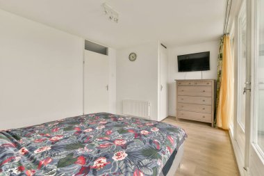 Rahat, modern bir yatak odası çiçek desenli bir yatak odası, ahşap döşeme ve doğal ışığa izin veren büyük pencereler, bir şifonyer ve duvar saati..