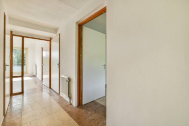 Geniş bir koridorda açık renkli duvarlar ve ahşap kapılar var. Doğal ışıkla aydınlatılıyor ve uzayda sıcak bir ortam yaratıyor..