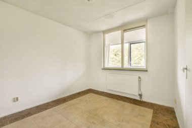 Parlak ve havadar bir atmosfere sahip olan minimalist oda, beyaz bir renk şeması, büyük bir pencere ve basit bir döşeme sergiliyor..