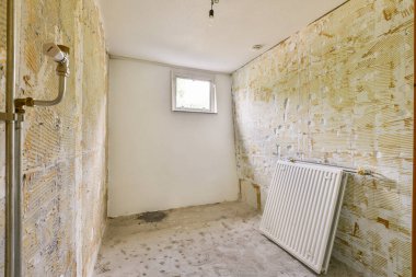 Boş bir odada çıplak duvarlar, doğal ışığa izin veren bir pencere ve duvara yaslanmış bir radyatör, minimalist bir atmosfer yaratıyor..