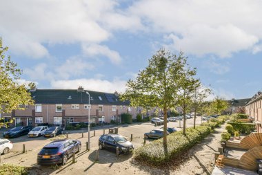 Parlak bir gökyüzünün altında ağaçların, park edilmiş arabaların ve tuğlaların olduğu sessiz bir yerleşim yeri. Sahne huzurlu bir şehir ortamı yakalıyor..