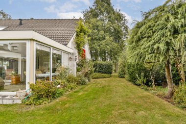 Yeşillik geniş pencereli modern bir evi çevrelemektedir. İyi korunan bahçede çimenler ve bitkiler var. Açık havada davet edici bir alan yaratıyorlar..