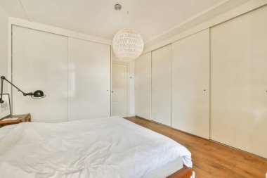 Beyaz bir yatak örtüsü, ahşap bir zemin ve çağdaş bir yaşam alanı için mükemmel, gösterişli dolap kapıları olan geniş ve havadar bir yatak odası..