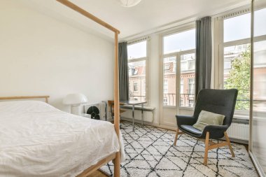 Parlak, havadar bir yatak odası, ahşap bir yatak odası, modern bir koltuk, ve büyük pencerelerden akan doğal ışık, huzurlu bir atmosfer yaratıyor..