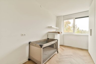 Parlak, modern bir çocuk odası, taşınabilir bir beşik ve camdan süzülen doğal ışıkla odaklanmış parlak bir masa içerir..