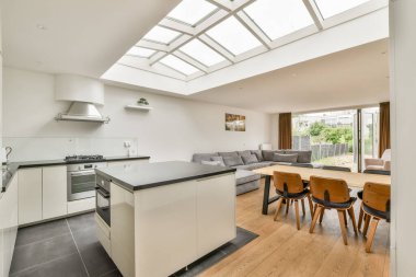 Zarif aletlerle donatılmış modern bir mutfak, geniş pencerelerden gelen doğal ışıkla dolu, rahat bir oturma odasına açılan açık bir düzen..