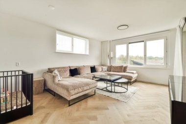 Modern bir oturma odasında şık bir kanepe, sehpa ve doğal ışık için geniş pencereler sergileniyor. Dekor çok az, rahat bir atmosfer yaratıyor..