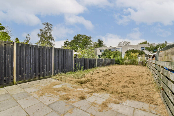 A serene, untended garden is surrounded by wooden fencing, creating a peaceful outdoor space. The area has patches of overgrown grass and sparse vegetation.