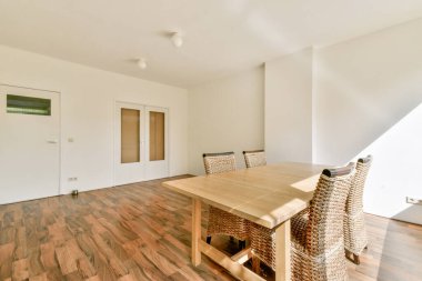Parlak, minimalist bir iç mekan ahşap bir yemek masası sergiliyor. Etrafı rattan sandalyelerle çevrili. Doğal ışık tasarımın basitliğini arttırır..