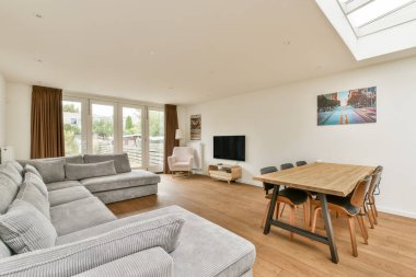 Geniş, modern bir oturma odası yumuşak mobilyalar, ahşap döşemeler ve geniş pencerelerden içeri akan bol miktarda doğal ışık..