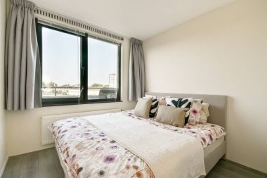 Geniş bir pencereden bol miktarda doğal ışıkla aydınlatılmış, çiçekli yatak ve dekoratif yastıklarla süslenmiş, rahat, modern bir yatak odası..