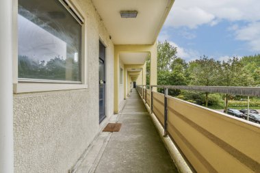 Modern bir apartman koridorunun aşağısında büyük pencerelerden yeşillik görünen bir manzara. Uzay temiz çizgiler ve doğal ışık ile tasarlanmıştır..