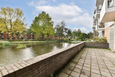 Arka planda ağaçlarla ve yerleşim yerleriyle dolu bir nehir kenarı yolunu gösteren sakin bir manzara, kentsel bir ortamda doğanın özünü yakalıyor..