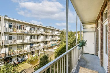 Yakın binaların ve açık mavi gökyüzünün yer aldığı modern bir apartman balkonundan çarpıcı bir manzara, konut mimarisi ve yeşilliği vurguluyor..