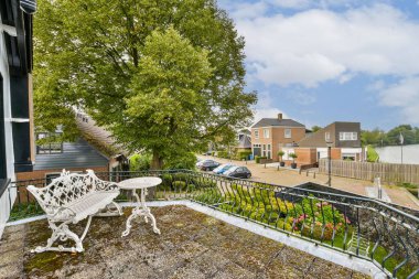 Resimli bir balkonda klasik mobilya ve bereketli yeşillik, sakin bir mahalle manzarasına bakıyor. Sahne, doğada huzurlu bir yaşam tarzını somutlaştırıyor..