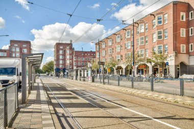 Hareketli bir şehirdeki tramvay istasyonunu gösteren canlı bir şehir manzarası. Modern binalar cadde boyunca uzanır, bulutlar mavi gökyüzünü benekler..