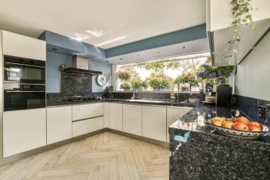 Zarif, modern bir mutfak, parlak bir dolap, mermer bir tezgah ve bol güneş ışığı alan büyük pencereler. Bir kase taze meyve, canlı atmosferi güçlendirir..