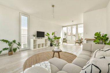 Geniş, modern bir oturma odası, hafif renk paleti, şık mobilyalar, ve kapalı bitkiler, dinlenmek için sakin bir atmosfer yaratıyor..