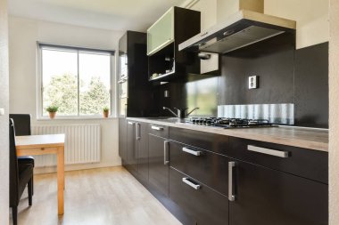 Koyu renk dolaplar, gaz sobası ve geniş bir pencereden yeşil manzaralı şık ve işlevsel bir alan yaratan şık ve modern bir mutfak..