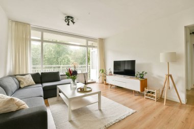 Rahat gri bir kanepe, ahşap mobilyalar ve geniş pencereli, doğal ışığın boşluğu doldurmasına olanak sağlayan modern bir oturma odası..