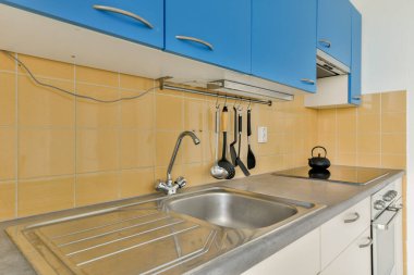 Mavi dolaplar, paslanmaz çelik lavabo, mutfak aletleri ve modern tasarım elementlerinin sergilendiği gösterişli bir düzenle modern bir mutfak..