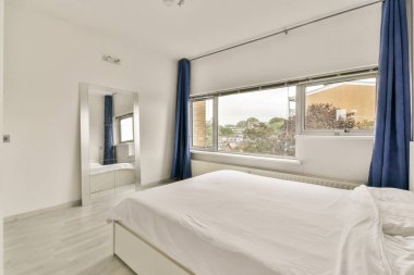 Geniş pencereleri, zarif perdeleri ve odanın ferah havasını arttıran minimalist bir tasarımı olan parlak ve çağdaş bir yatak odası..
