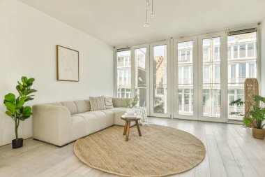 Aydınlık bir kanepe, yuvarlak bir halı ve doğal ışığı davet eden büyük pencereli parlak ve havadar bir oturma odası. Kapalı alandaki bitkiler modern mekana biraz doğa katar..
