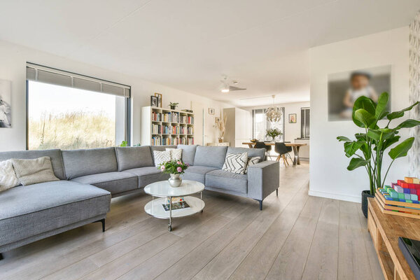 Стильная современная гостиная с серым диваном, стеклянным журнальным столиком и большими окнами с естественным освещением с акцентом на зеленые растения.