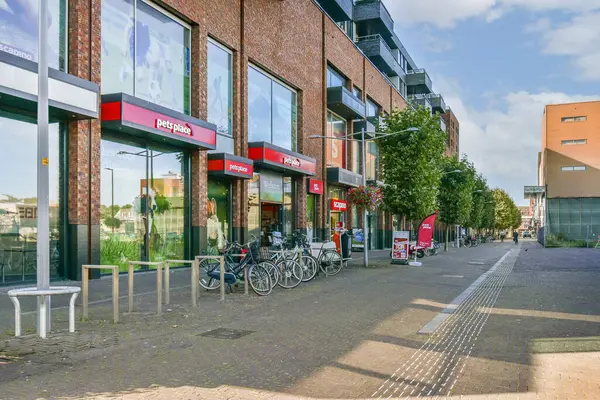 Canlı bir sokak sahnesi modern perakende satış yerleri, park edilmiş bisikletler ve kaldırımı çevreleyen yeşillik, canlı bir şehir atmosferini gözler önüne seriyor..