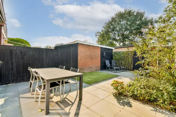 Çağdaş bir açık hava verandası gösterişli bir masa ve yemyeşil bitkilerle çevrili sandalyeler ve arka planda açık mavi bir gökyüzü..