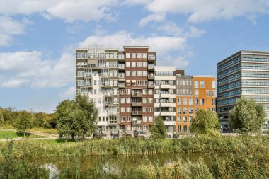 Modern apartman binaları sakin bir parkın yanında duruyor, mimari çeşitlilik ve suya yakın yeşil alan entegrasyonu sergileniyor.