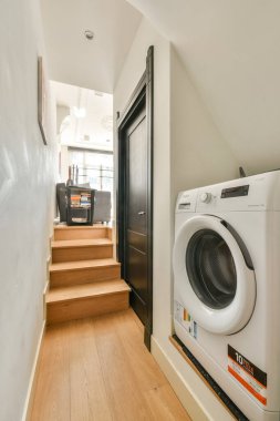 Ahşap merdivenlerin yanında çamaşır makinesinin bulunduğu şık bir çamaşır odası, işlevselliği çağdaş tasarımla harmanlıyor..