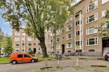 Görüntü, yeşil ve park edilmiş araçlarla çevrili modern bir şehir apartmanını gösteriyor. Geniş pencereler ve hoş bir atmosferi var..