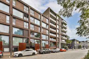 Görüntü, yakınındaki geniş pencereli ve yeşillikli çağdaş bir apartmanı bulutlu bir gökyüzüne karşı sergiliyor. Şık bir şehir ortamı tasvir edilir.