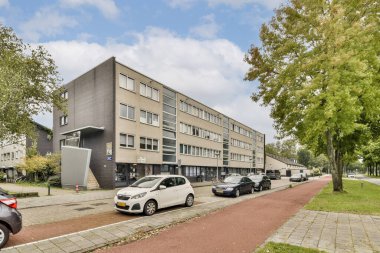 Çağdaş bir mimari tasarım, şehir caddesinde bakımlı yeşillik ve park halindeki arabaların yanında modern bir bina sergiliyor..