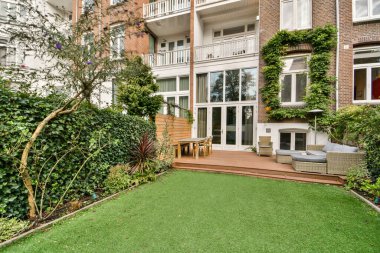 Bereketli bir yeşillik, ahşap bir veranda ve şık bitkiler içeren çağdaş bir açık alan, şehir ortamında rahatlama ve açık hava buluşmaları için idealdir..