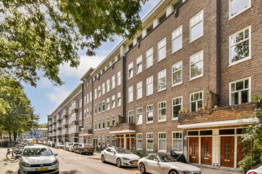 Canlı bir şehir manzarası açık mavi gökyüzünün altında yemyeşil ve park halindeki arabalarla çevrili bir dizi modern bina sergiliyor..