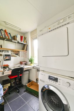 Masa, kitaplık ve çamaşır makinesinin yer aldığı, işlevsel çift kullanımlı bir alanı gösteren iyi organize edilmiş modern bir ev ofisi..