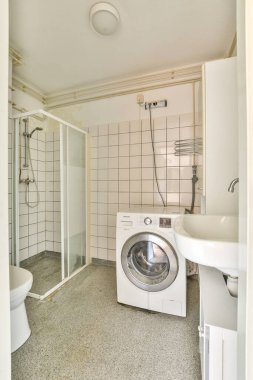 Gösterişli ve minimalist bir banyo. İçinde çamaşır makinesi, duş alanı ve temiz hatlar var. Parlak alan işlevselliği ve modern tasarımı vurgular.