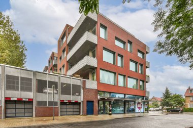 Bu resim, geniş pencereli ve parlak dizaynlı, gür ağaçlar ve açık gökyüzüyle çevrili çağdaş bir binayı gösteriyor, kentsel yaşamı vurguluyor..