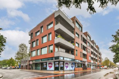 Geniş pencere ve balkonların yer aldığı çarpıcı bir modern bina tasarımı, merkezdeki canlı perakende işaretiyle tamamlanmış, kentsel bir çevrede yer alıyor..