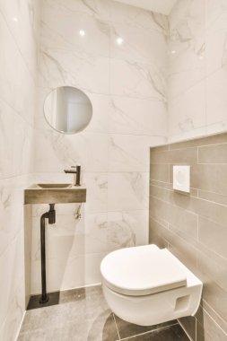 Duvarlarında mermer fayanslar olan küçük bir banyoda beyaz bir tuvalet ve üzerinde yuvarlak bir ayna asılı.