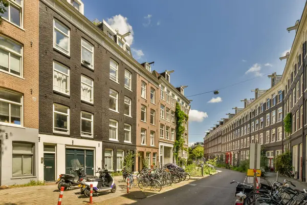 Bazı motosikletler sokak kenarına park edilmiş. Şehir merkezinde binalar ve sokakta yürüyen insanlar var.