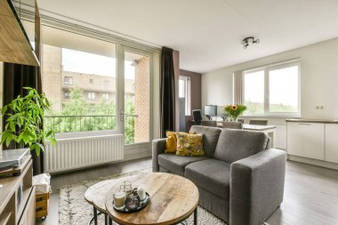 Rahat bir kanepe, ahşap bir sehpa ve geniş pencerelerden yeşillik manzaralı, parlak ve havadar modern bir oturma odası. Hoş bir atmosfer yaratıyor..