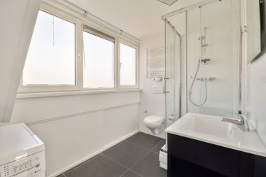 Cam duş, tuvalet ve çamaşır makinesinin yer aldığı gösterişli, modern bir banyo tasarımı. Geniş pencerelerden gelen geniş doğal ışıkla vurgulanmış..