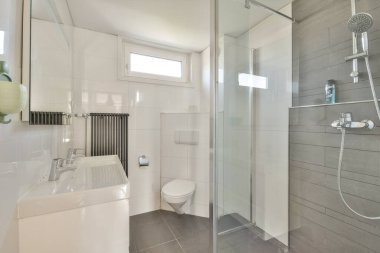 Gösterişli, çağdaş bir banyo, cam duş muhafazası, minimalist lavabo tasarımı, ve nötr fayans renkleri, modern estetik ve işlevsellik sergileniyor..