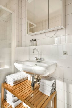 Ahşap bir raf, beyaz bir lavabo, bolca havlu ve çağdaş tasarım elementlerinin bulunduğu şık bir banyo..