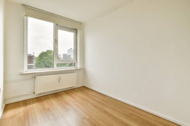 Geniş pencereli, şehir manzarası ve dışarıdaki yeşilliği gösteren ferah bir minimalist oda. Ahşap zemin iç kısmın basitliğini arttırır..